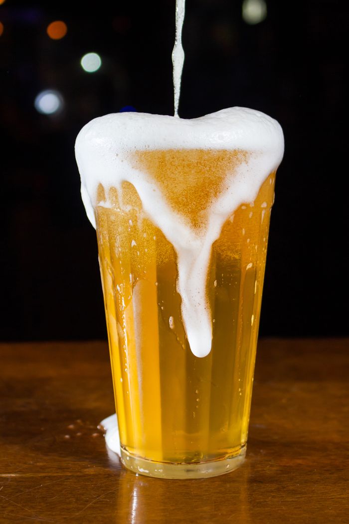 Close-up of a tall beer glass overflowing with foam on a wooden bar table at night.